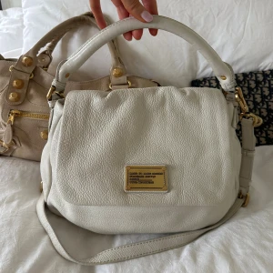 Vit Lil Ukita bag med gulddetaljer - Snygg och praktisk Marc by Marc Jacobs Classic Q Lil Ukita i mjukt vitt pebble-läder med guldfärgade detaljer. Väskan har lock och top handle så att den kan bäras på armen. Perfekt storlek för varda  Inuti finns dragkedjefack och öppna fack för enkel organisering. En populär modell ur Classic Q-serien och en eftertraktad vintage-väska.  Mått ca 33×26×9 cm. Kan hämtas i Nacka eller skickas 🤍Skriv för fler bilder!