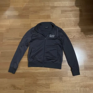 Grå EA7 track jacket med dragkedja - Snygg grå track jacket från EA7 Emporio Armani med vit logga på bröstet och diskret detalj på ryggen. Jackan har hel dragkedja, ribbade muddar och två fickor framtill. Perfekt för en sportig och stilren look.