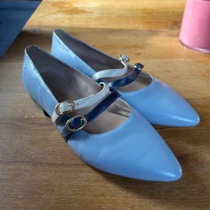 Blå ballerinaskor med spännen från Bagatt - Snygga ljusblå ballerinaskor i skinn från Bagatt med spetsig tå och två remmar över foten, en i krämvit och en i mörkblått, båda med guldfärgade spännen. Aldrig andvända.