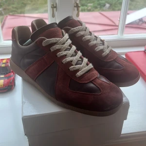 Vin Röda Maison margela  - Snygga maison sneakers i mörkbrunt skinn med matchande mockadetaljer och beige insida. Skorna har vita snören och en gummisula med coolt mönster undertill. Perfekt för dig som gillar retrovibbar och vill sticka ut med en unik look.