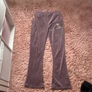  Lila velourbyxor från Juicy Couture - Säljer ett par lila velourbyxor från Juicy Couture med bootcut-ben och snörning i midjan. Byxorna har en glittrig Juicy Couture-logga med strass på framsidan och en bakficka. Perfekta för en chill och trendig look.