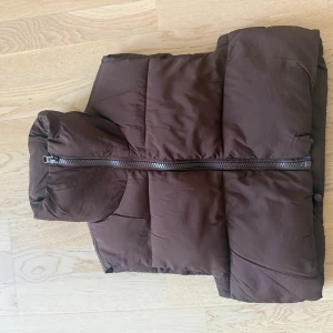 Brun dunväst med huva - Snygg brun dunväst med stor huva och grov dragkedja framtill. Västen är quiltad och har en puffig look som ger en trendig siluett. Perfekt att slänga över en hoodie för extra värme under kyliga dagar.