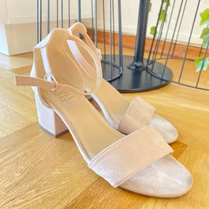 Beige sandaler med klack från Bullboxer - Snygga sandaler från Bullboxer i ljusbeige mocka med bred rem över tårna och justerbar rem runt ankeln. Skorna har öppen tå, blockklack och en mjuk, lyxig känsla. Perfekta för sommarens alla tillfällen när du vill känna dig extra fin.