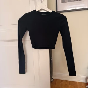 Svart croppad långärmad topp Zara - En svart, croppad långärmad topp från Zara. Enkel och stilren design med rund halsringning och tight passform. Tillverkad i mjuk bomull som känns skön mot huden. Perfekt att matcha med höga jeans eller kjol för en clean look.