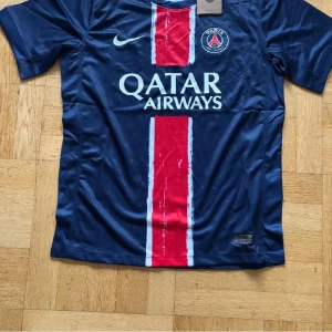 PSG matchtröja Nike blå/röd - Snygg PSG fotbollströja från Nike i marinblått med röd vertikal rand och vita detaljer. Tröjan har klubbmärke på bröstet, Qatar Airways-tryck och klassisk passform. Perfekt för dig som älskar fotboll och vill visa ditt stöd för Paris Saint-Germain.