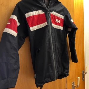 Svart vindjacka Helly Hansen - Svart Helly Hansen Jackan har huva, hel dragkedja framtill och justerbar nederkant. Ett resårband trasigt nederkant 