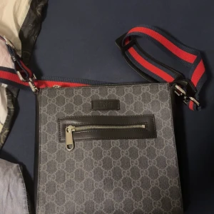 Svart axelväska från Gucci med GG-mönster - Snygg svart axelväska från Gucci med klassiskt GG-mönster över hela väskan. Väskan har en framficka med dragkedja och justerbar axelrem i blått och rött. Insidan har flera fack och detaljer i svart skinn. Perfekt för dig som vill ha en stilren och lyxig väska.