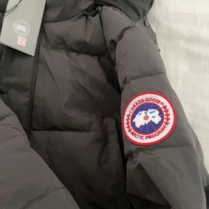 Svart dunjacka från Canada Goose - Svart dunjacka från Canada Goose med ikonisk patch på ärmen. Jackan har huva, quiltad design och dragkedja framtill. Perfekt för kalla vinterdagar och har en clean, stilren look som funkar till allt. Klassisk modell med lång ärm och snygga detaljer.