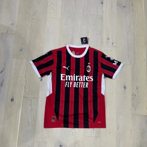 AC Milan kortärmad fotbollströja Puma - Säljer en officiell AC Milan fotbollströja från Puma med klassiska röda och svarta vertikala ränder, vit krage och vita detaljer på ärmarna. Tröjan har klubbmärke och sponsortryck på bröstet. Tillverkad i lätt och ventilerande polyester.