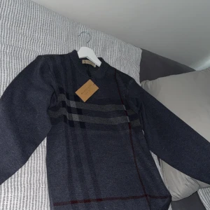 Burberry Tröja - Snygg mörkgrå tröja från Burberry med klassiskt rutmönster i grått och vinrött. Tröjan har långärmad passform och rund halsringning, perfekt för dig som gillar stilrena och tidlösa plagg. Materialet känns mjukt och skönt mot huden.