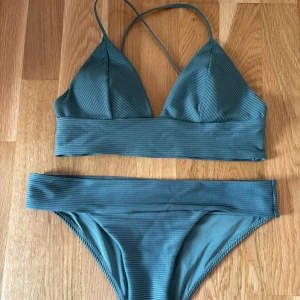 Ribbad olivgrön bikini - Snygg bikini i mörkgrön olivgrön ish färg med ribbad struktur. Toppen har trekantsformade kupor och smala axelband som korsas i ryggen med guldiga detaljer. Underdelen har en bred linning som ger en cool och modern look. Perfekt för dig som gillar stilrena detaljer och trendig design. Passar str 34-38
