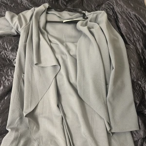 Ljusgrå omlottkappa med knytband - Säljer en ljusgrå kappa i omlottmodell med knytband i midjan. Jackan har långa ärmar och ett mjukt, lätt tyg som ger ett snyggt fall. Perfekt för dig som gillar stilrena och enkla plagg med en modern touch.