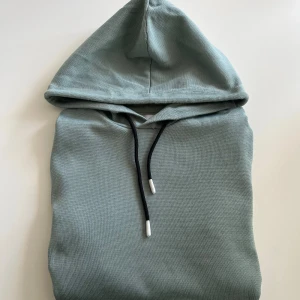 Ljusgrön hoodie från Jack & Jones - Snygg ljusgrön hoodie från Jack & Jones med svart snörning i huvan och ribbade muddar. Tröjan är i mjuk bomull och har lång ärm samt en diskret logga på ärmen. Perfekt för chill dagar och enkel att matcha med jeans eller joggers.