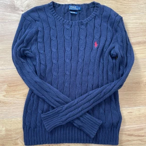 Mörkblå kabelstickad tröja Polo Ralph Lauren - Kabelstickad mörkblå tröja från Polo Ralph Lauren i mjuk pima bomull. Klassisk rund halsringning, långärmad modell och ikonisk röd broderad logga på bröstet. Skicket är 10/10