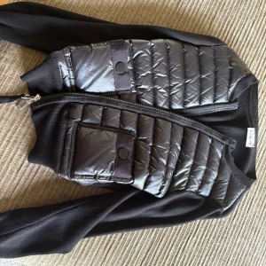 Svart puffer cardigan jacka Moncler - Snygg svart cardigan jacka från Moncler med puffer-detaljer framtill och släta ärmar. Jackan har dragkedja, rund halsringning och en tight passform. Perfekt för dig som vill ha en sportig men ändå stilren look.