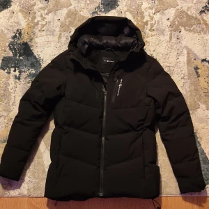 Svart dunjacka från Sail Racing storlek S - Svart dunjacka från Sail Racing med huva och dragkedja framtill. Jackan är vadderad med 500 FP dun från Allied Feather + Down och har en clean, sportig look. Perfekt för kalla dagar och har en snygg logga på bröstet och huvan.