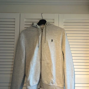 Ljusgrå hoodie från Polo Ralph Lauren - Snygg ljusgrå hoodie från Polo Ralph Lauren med broderad logga på bröstet. Tröjan har huva med snörning, dragkedja framtill och två stora fickor. Perfekt för chill dagar och enkel att matcha med jeans eller joggers. Köpt på pondus mycket bra skick mvh !