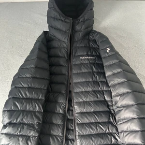 Svart dunjacka Peak Performance - Svart dunjacka från Peak Performance med huva och horisontella sömmar. Jackan har dragkedja framtill, innerfickor och diskret logga på bröstet och ärmen. Perfekt för kyliga dagar och riktigt lätt att bära.