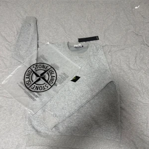 Grå sweatshirt från Stone Island - Säljer en stilren grå sweatshirt från Stone Island i bomull. Tröjan har rund hals, ribbade muddar och den klassiska loggan på vänster ärm. Perfekt för dig som gillar clean streetwear och vill ha något bekvämt och snyggt. Det står att storleken på denna varann ör M men den passar perfekt på mig, och jag använder S. Skriv till mig innan ni köper.