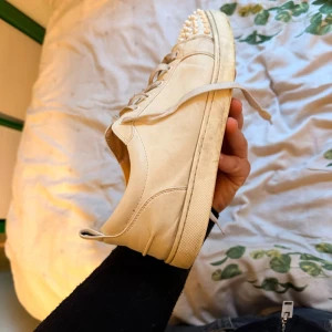 Vits Christian Louboutin sneakers med nitar - Säljer ett par beiga sneakers från Christian Louboutin med ikonisk röd sula och coola nitar framtill. Skorna är i skinn och har snörning. Kommer med originalpåse och box. Perfekta för dig som vill sticka ut med en lyxig och edgy vibe.