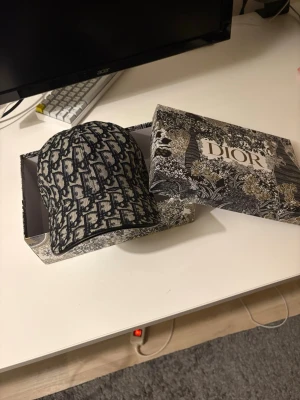 Dior monogram keps i bomull - Säljer en exklusiv Dior keps med ikoniskt monogrammönster i svart och grått. Kepsen är tillverkad i 100% bomull och har justerbar rem bak. Kommer i originalförpackning med snyggt mönstrad Dior-box. Perfekt för dig som vill ha en lyxig och trendig accessoar.