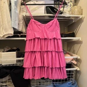 Rosa volangklänning med smala axelband - Säljer en rosa kortklänning med smala axelband och flera lager volanger. Klänningen har en feminin siluett med markerad byst och är tillverkad i ett lätt, plisserat tyg som ger ett luftigt intryck. Perfekt för varma dagar eller sommarens tillställningar.