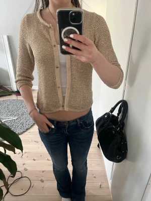 Guld kofta med knappar - Snygg stickad kofta med rund hals och knäppning framtill. Koftan har trekvartsärmar och ett luftigt, strukturerat stickmönster. 