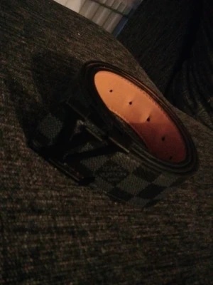 Louis Vuitton rutigt bälte svart/grå - Snyggt bälte från Louis Vuitton med klassiskt rutigt mönster i svart och grått. Bältet har en svart LV-logga som spänne och insidan är i ljusbrunt läder. Perfekt accessoar för att lyfta din outfit.