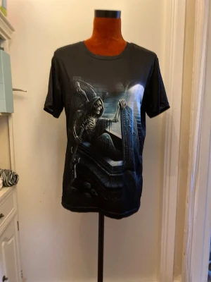Svart t-shirt med dödskalletryck S - Cool svart t-shirt med stort tryck av en skelettfigur som reser sig ur en grav, med detaljer som gravsten och månsken. Perfekt för dig som gillar mörka och unika motiv. Rund halsringning och klassisk passform.