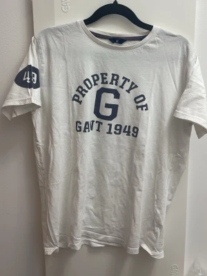 Vit t-shirt från Gant med tryck - Vit t-shirt från Gant med blått tryck 'PROPERTY OF G GANT 1949' på bröstet och siffran 49 på ärmen. Klassisk rund hals och korta ärmar. Perfekt för en avslappnad och sportig stil.