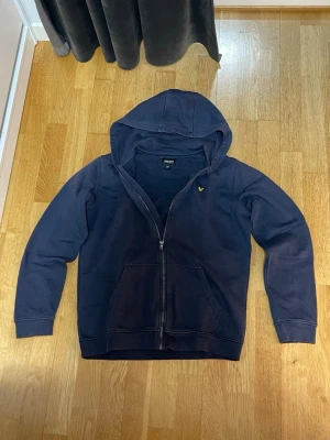 Mörkblå hoodie från Lyle & Scott - Mörkblå hoodie från Lyle & Scott med dragkedja framtill och klassisk gul logga på bröstet. Tillverkad i mjuk bomullsblandning och har två fickor fram samt en stor huva. Perfekt för en chill och avslappnad stil.