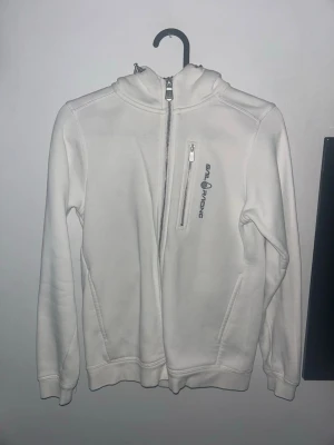 Vit hoodie från Sail Racing - Säljer en stilren vit hoodie från Sail Racing med dragkedja snett över bröstet och en liten bröstficka med logga. Hoodien har huva och ribbade muddar vid ärmar och nederkant. Perfekt för dig som gillar sportig och clean stil.