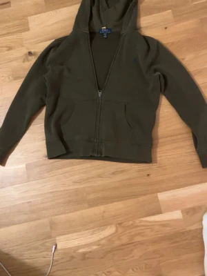 Olivgrön hoodie från Polo Ralph Lauren - Snygg olivgrön hoodie med dragkedja från Polo Ralph Lauren. Klassisk modell med huva, två fickor framtill och diskret logga på bröstet. Perfekt för en avslappnad streetwear-look. Mjuk bomullsblandning som känns skön mot huden. Passar ungefär nån som är 160 kan sänka vid snabb affär hör av er om de är något 
