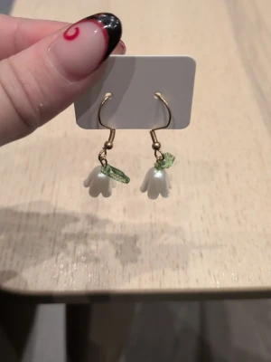 Blommiga örhängen med blad i rostfritt stål  - Säljer ett par söta örhängen med vita blommor och gröna blad i akryl. Örhängena har guldfärgade krokar och en lekfull, trendig design som passar perfekt till dig som gillar unika accessoarer. Perfekt detalj för att lyfta din outfit!