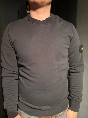 Svart sweatshirt från Stone Island - Säljer en svart sweatshirt från Stone Island med den klassiska logotypen på vänster ärm. Tröjan har rund halsringning, ribbade muddar och är tillverkad i mjukt bomullsmaterial. Perfekt för en avslappnad och stilren look. Tröjan är äkta och i helt nytt skick.