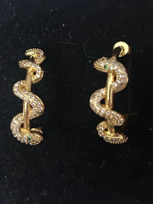Guldiga ormörhängen med stenar - Säljer ett par unika örhängen i form av ormar, tillverkade i guldfärgad metall och dekorerade med små gnistrande stenar. Ormarnas ögon har gröna detaljer som ger extra edge. Perfekta hoops för dig som vill sticka ut och addera lite attityd till din look. Endast testade! Box ingår!