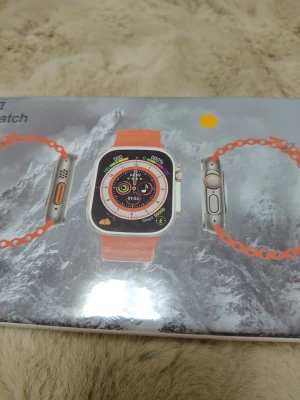 Apple Watch Ultra II med orange band - Säljer en Apple Watch Ultra II med ett snyggt orange silikonband. Klockan har en stor, fyrkantig skärm med silverfärgad ram och flera smarta funktioner för träning och hälsa. Perfekt för dig som vill ha koll på både aktivitet och notiser direkt på handleden.