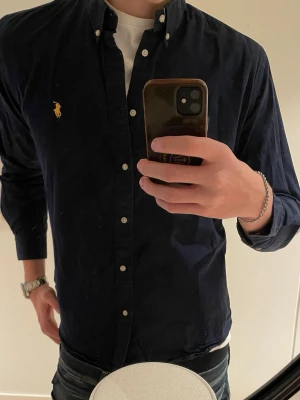 Mörkblå skjorta från Ralph Lauren - Klassisk mörkblå skjorta från Ralph Lauren med knappkrage och den ikoniska gula logon broderad på bröstet. Skjortan har långa ärmar och vita knappar framtill. Perfekt till jeans eller chinos för en stilren look.