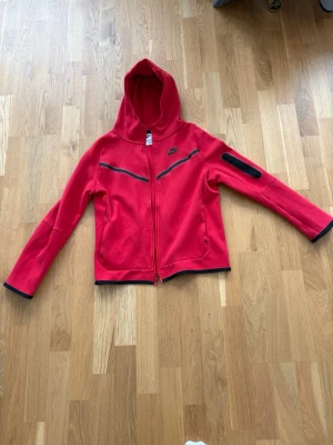 Röd Nike tech fleece - Nike hoodie i rött i storlek 147-158 med hel dragkedja och svart logga på bröstet. Två fickor framtill och en extra ficka med dragkedja på ena ärmen. Svarta detaljer längs dragkedja, ärmslut och nederkant. Tillverkad i mjukt bomullsmaterial och har en stor huva.