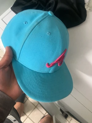 Turkos New Era 59FIFTY keps - Säljer en turkos New Era 59FIFTY keps med röd broderad 'A' framtill. Klassisk flat brim och strukturerad passform. Märkt med MLB och officiell merchandise. Perfekt för dig som vill sticka ut med färg och stil.