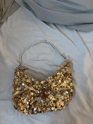 Guldig paljett handväska - Säljer en superglittrig handväska täckt av guldiga paljetter. Väskan har en kort, silvrig kedjerem som ger en lyxig känsla. Perfekt för dig som vill sticka ut och addera lite extra bling till din outfit.
