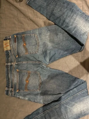 Blå Nudie Jeans med raka ben - Säljer ett par klassiska blå jeans från Nudie Jeans med snygga slitningar och slimfit passform.