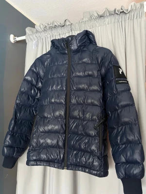 Mörkblå dunjacka Peak Performance - Snygg mörkblå dunjacka från Peak Performance med huva och dragkedja framtill. Jackan har två sidofickor med dragkedja och en logga på armen. En av fickorna går inte att stänga med dragkedjan. Bara att fråga frågor om det är nåt.