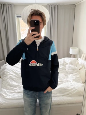 Blå vindjacka från Ellesse - Säljer en snygg vindjacka från Ellesse i marinblått med ljusblå och vita detaljer. Jackan har huva med snörning, stor logga framtill och halv dragkedja. Perfekt för dig som gillar sportig stil och vill ha något lätt och bekvämt.
