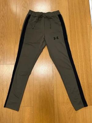 Svart Under Armour byxor  - Snygga olivgröna träningsbyxor från Under Armour med svarta sidopaneler och dragsko i midjan. Byxorna har en smal passform och är tillverkade i ett stretchigt funktionsmaterial som passar perfekt till träning eller chill. Size S men passar M och ksk L, pris lan diskuteras. Jag har aldrig använt de för de passar inte mig.