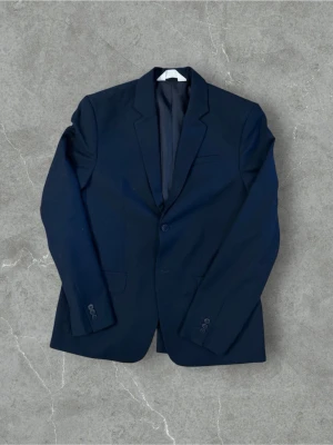 Mörkblå klassisk kavaj - Stilren mörkblå kavaj med två knappar framtill och klassiska slag. Kavajen har innerfoder och flera fickor, perfekt för en dressad look. Passar till både skjorta och t-shirt för en snygg outfit. Storlek Eu 164) men passar mig som är runt 180 skulle sitta perfekt på 175cm