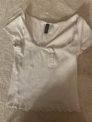 Vit ribbad topp från Gina Tricot - Snygg vit ribbad topp från Divided med korta ärmar och knappar framtill. Toppen har en lätt croppad passform, vågig kant nertill och vid ärmslut samt en djupare rundad halsringning.