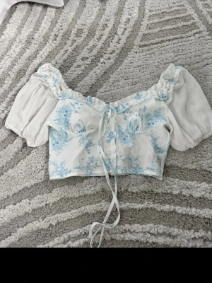 Vit blommig croppad blus med puffärm - Supersöt croppad blus i vitt med ljusblått blommönster och puffiga korta ärmar. Toppen har snörning framtill, dragkedja bak och volangkant vid urringningen. Perfekt för en romantisk look och riktigt trendig just nu.