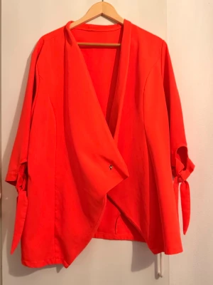 Orange omlottkofta med knytärmar - Snygg röd omlottkofta med trekvartsärmar och knytband vid ärmsluten. Koftan har en lös och avslappnad passform med öppen framsida, perfekt för lager på lager. Tillverkad i ett mjukt tyg som känns bekvämt mot huden.