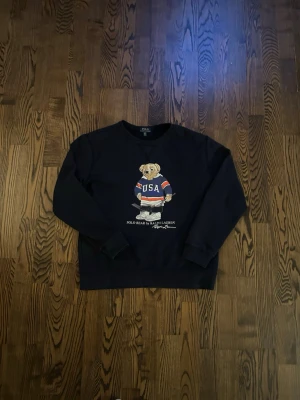 Mörkblå  stickad tröja Polo Bear Ralph Lauren - Mörkblå stickad tröja från Ralph Lauren med ikoniska Polo Bear-trycket på bröstet. Björnen har en blå tröja med 'USA' och håller en hockeyklubba. Tröjan har rund hals och långa ärmar. Perfekt för dig som gillar streetwear och sportiga vibes.